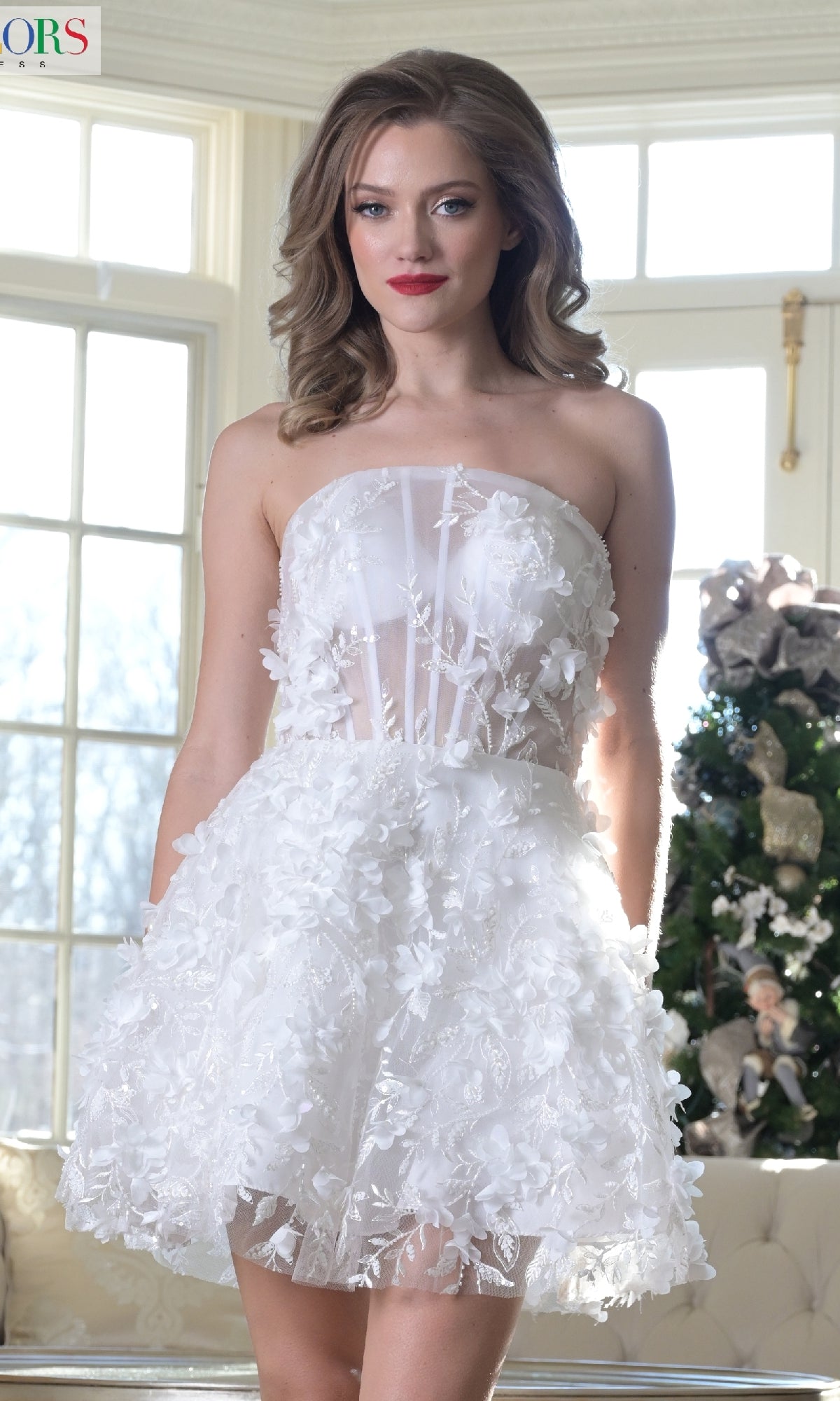 A woman wears the Strapless Floral Homecoming Dress: Colors Dress 3659, featuring 3-D flower embellishments, as she stands indoors by a large window and a decorated Christmas tree.
